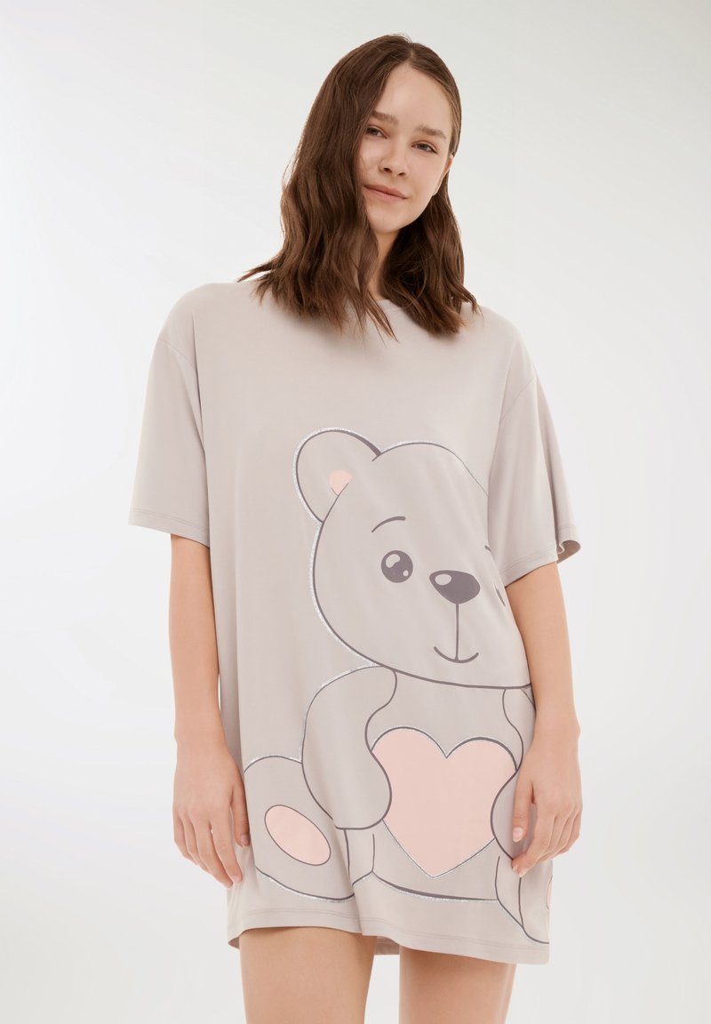 Giovane donna con capelli castani ondulati che indossa un abito beige oversize con un grande orsetto di peluche che tiene in mano un cuore rosa.