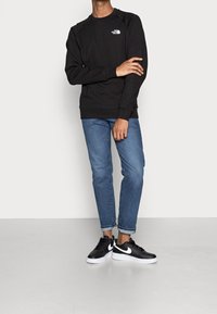 Svart sweatshirt med rund hals och vitt logo, kombinerad med ljusblå denimjeans och svarta sneakers med vit sula.