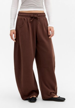 Unterer Torso mit lockeren braunen Hosen mit Kordelzug, weißem Langarmshirt und schwarzen flachen Schuhen vor weißem Hintergrund.