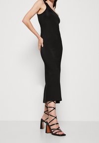 Kobieta w dopasowanej czarnej sukience midi na jedno ramię oraz czarnych sandałach na wysokim obcasie i w paski, stojąca na tle białym.