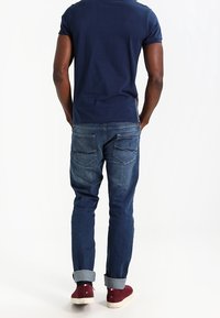 Granatowa koszulka w połączeniu z dżinsami z podwiniętymi nogawkami. Dżinsy są ciemnoniebieskie z wyblakłym wykończeniem i mają dwie tylne kieszenie. Na nogach czerwone trampki.