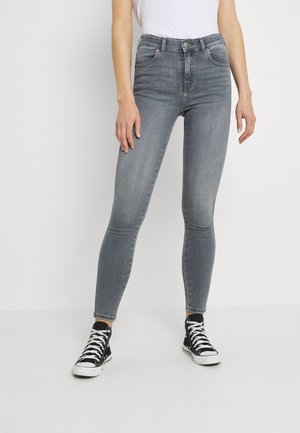 Persoon draagt hooggesneden grijze skinny jeans, wit shirt en zwarte hoge sneakers, staand tegen een witte muur.