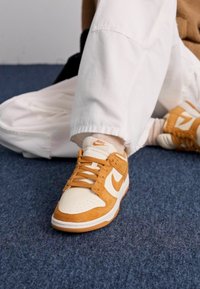 Nike Dunk tossud, millel on koorekarva nahaküüs, tan värvi suede detailid, aukudega pind ja oranž bränding. Taustal on sinine vaip.