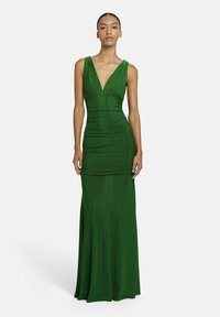 Eine Frau steht vor einem schlichten Hintergrund und tr�ägt ein ärmelloses, tief ausgeschnittenes, bodenlanges, funkelndes grünes Abendkleid.