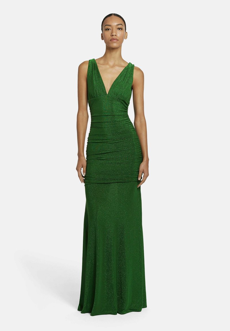 Eine Frau steht vor einem schlichten Hintergrund und trägt ein ärmelloses, tief ausgeschnittenes, bodenlanges, funkelndes grünes Abendkleid.