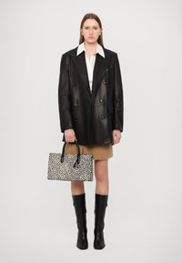 Blazer noir en cuir à double boutonnage, chemise blanche, jupe brune, bottes noires à genoux et un sac blanc avec des taches noires, tenu à la main.