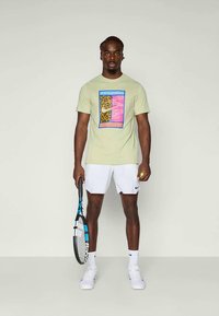 Svijetlozeleni pamučni majica s šarenim grafičkim dizajnom koji uključuje uzorak leoparda i geometrijske oblike. Nosite s bijelim sportskim kratkim hlačama.