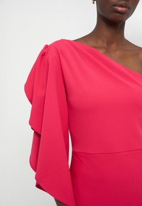 Femme portant une robe rose vif à épaules dénudées avec une grande manche drapée sur le côté gauche, sur un fond uni.