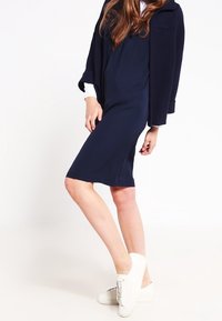 Žena v námořnickém šatu, námořnické zkrácené bundě, bílé košili a bílých teniskách stojí proti jednobarevnému bílému pozadí.