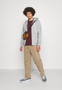 Grauer Zip-Hoodie, kastanienbraunes T-Shirt, beige Hose, graue Sneaker mit weißen Sohlen und eine kleine braune Umhängetasche. Lockige Haare, stehend.