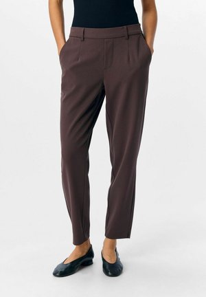 Person trägt schmal geschnittene, dunkelbraune Hose mit Seitentaschen und schwarze Schuhe mit niedrigem Absatz, steht vor einem einfarbigen Hintergrund.