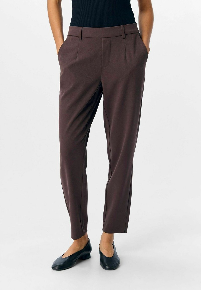 Personne portant un pantalon brun foncé ajusté avec poches latérales et des chaussures noires à talons bas, debout devant un fond uni.