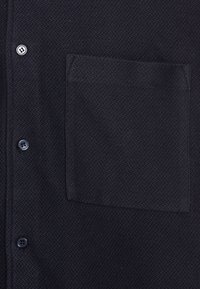 Camicia nera con motivo a spina di pesce testurizzato, chiusura con bottoni e una tasca applicata sul lato sinistro.