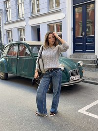 Pelēks džemperis ar rakstu adījumu, platām zilām džinsa biksēm un brūnām kurpēm; fonā vintāžas zaļš auto; pilsētas ielas ainava ar pasteļtoņu ēkām.