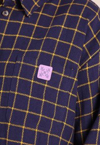 Chemise tricotée bleu marine avec un motif à carreaux jaunes, bouton marron près du col et patch carré rose avec un motif floral sur la poche poitrine.