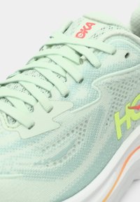 Ανοιχτό πράσινο αθλητικό παπούτσι Hoka με λεπτομερή πλέγμα στο πάνω μέρος, ανοιχτό πράσινο κορδόνια και κίτρινο λογότυπο στη λευκή και πορτοκαλί σόλα.
