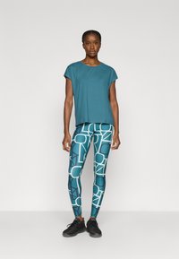 Τιρκουάζ τοπ με κοντά μανίκια και τιρκουάζ κολάν με αφηρημένα λευκά γεωμετρικά σχέδια. Το μοντέλο φοράει μαύρα αθλητικά παπούτσια.