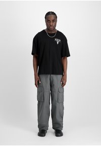 T-shirt noire surdimensionnée avec le texte "BERLIN", pantalon cargo gris avec poches et chaussures noires à enfiler. Modèle se tient contre un fond blanc.