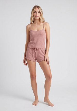 Kvinde stående barfodet iført en lyngfarvet ribbet tanktop og matchende shorts mod en ensfarvet lys grå baggrund.