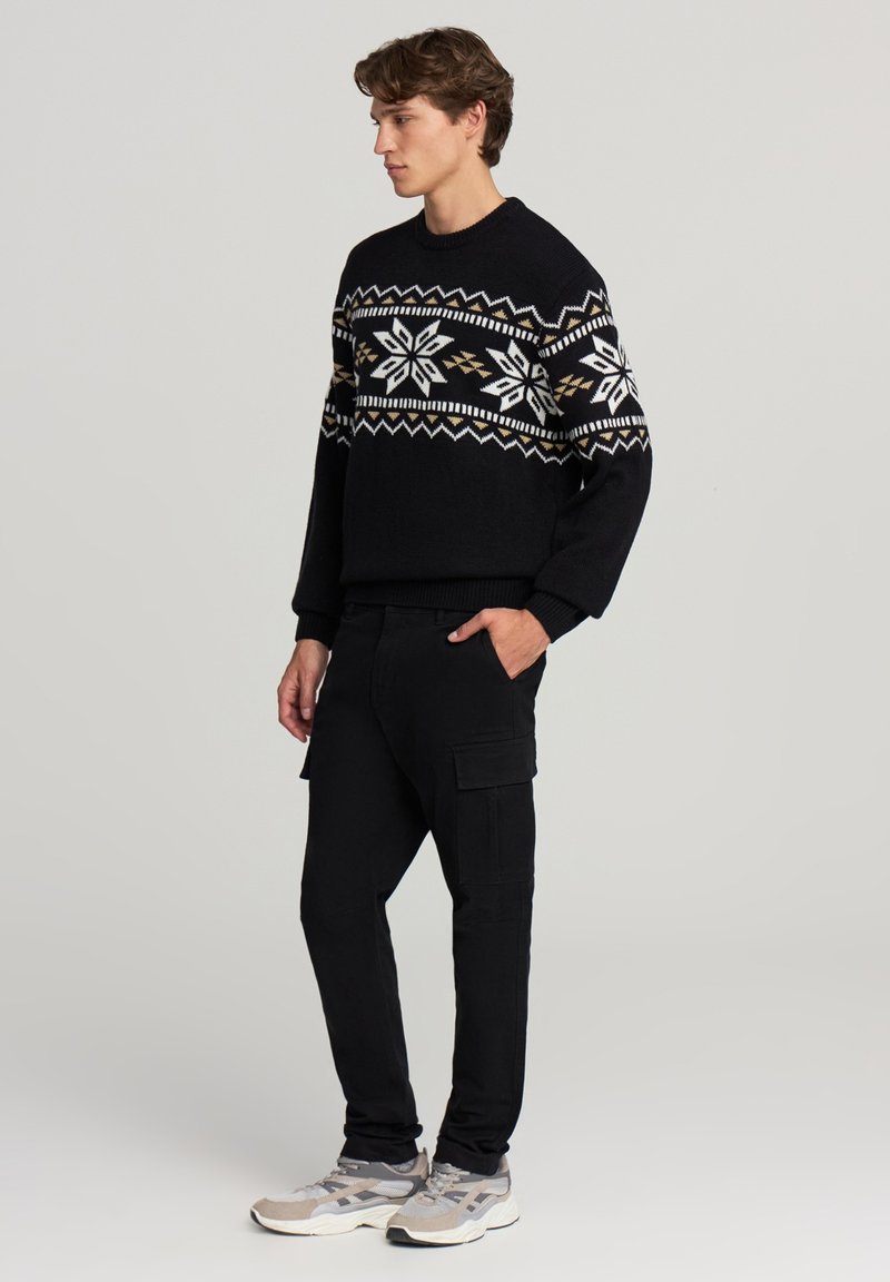 Czarny sweter dzianinowy z białymi i złotymi wzorami geometrycznymi, zestawiony z czarnymi spodniami cargo. Model nosi szare i czarne buty sportowe.