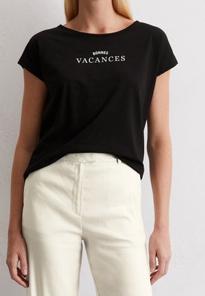Kvinde iført sort T-shirt med teksten "BONNES VACANCES" og hvide højtaljede bukser, stående mod en ensfarvet lys væg.