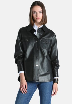 Femme aux cheveux châtains mi-longs porte une veste en cuir noire boutonnée sur une chemise blanche et un jean bleu sur un fond uni.