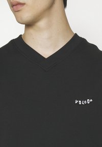 Gros plan d'une personne portant un t-shirt noir à col en V avec un petit logo blanc « Volcom » sur la poitrine, tête partiellement visible regardant vers la gauche.