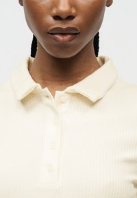 Κοντινό πλάνο ενός ατόμου που φοράει πλεκτό πόλο μπλουζάκι σε κρεμ χρώμα με ριμπ υφή, γιακά και τέσσερα κουμπιά, εστιάζοντας στο λαιμό και το πάνω μέρος του στήθους.