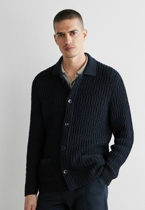 Man with short hair wearing a black textured button-up cardigan over a beige collared shirt, standing against a plain white wall.