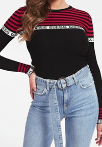 Musta ja punainen raidallinen pitkähiha topi, jossa on logonauha kaula-aukossa, yhdistettynä korkeavyötäröisiin vaaleansinsisiin farkkuihin ja pyöreään vyösolkeen.