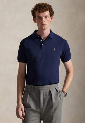 Jeune homme aux cheveux roux bouclés portant un polo bleu marine et un pantalon plissé gris, debout devant un fond beige uni.