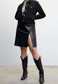 Giacca di jeans nera sopra una maglietta a righe bianca e nera, abbinata a una gonna nera in ecopelle con spacco laterale e stivali neri al ginocchio.