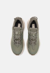 Zielone buty trekkingowe z siateczkowym wierzchem o teksturowanej fakturze, elastycznymi sznurowadłami, wyściełanym kołnierzem oraz grubą beżową podeszwą z czarnymi akcentami.