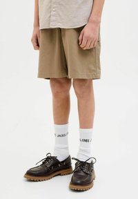 Shorts kaki en coton, design structuré, poche latérale. Chaussures marron à lacets avec semelles épaisses, chaussettes blanches à côtes avec texte noir.