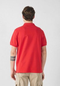 Rood poloshirt met een kraag en korte mouwen, gemaakt van gladde stof. De achterkant heeft een rechte zoom en een zijsplit voor extra draagcomfort.