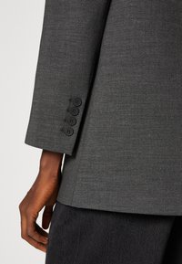 Donkergrijze blazer met een gladde textuur, voorzien van een enkele split en vier zwarte knopen op de mouw, gemaakt van een op maat gemaakte stof.
