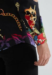 Chemise à manches longues en tissu noir ornée de motifs colorés de chaînes, de feuilles et de designs abstraits. Accents rouges aux poignets avec des boutons.