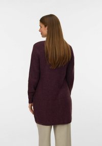 Pull en tricot bordeaux long avec une finition texturée, présentant une coupe droite et des fentes sur les côtés, porté sur un pantalon beige.