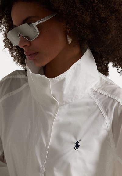 Weiße wasserdichte Jacke mit hohem Kragen, mit einem schwarzen gestickten Logo. Silberne verspiegelte Sonnenbrille mit "POLO"-Markenlogo, sichtbare strukturierte Locken.