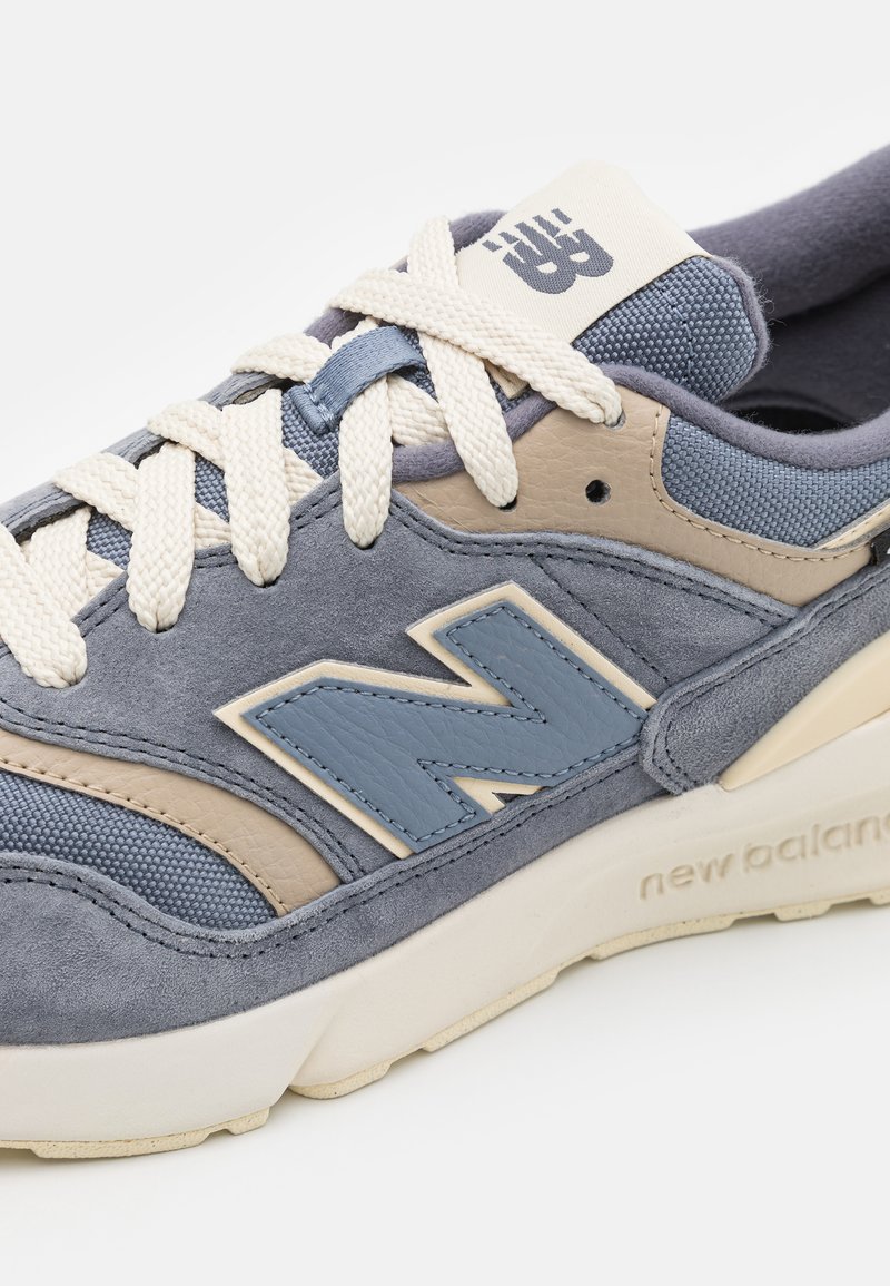Blaue und beige New Balance Sneaker mit weißen Schnürsenkeln, Obermaterial aus Wildleder und Mesh, auffälligem "N"-Logo und gepolsteter Sohle.