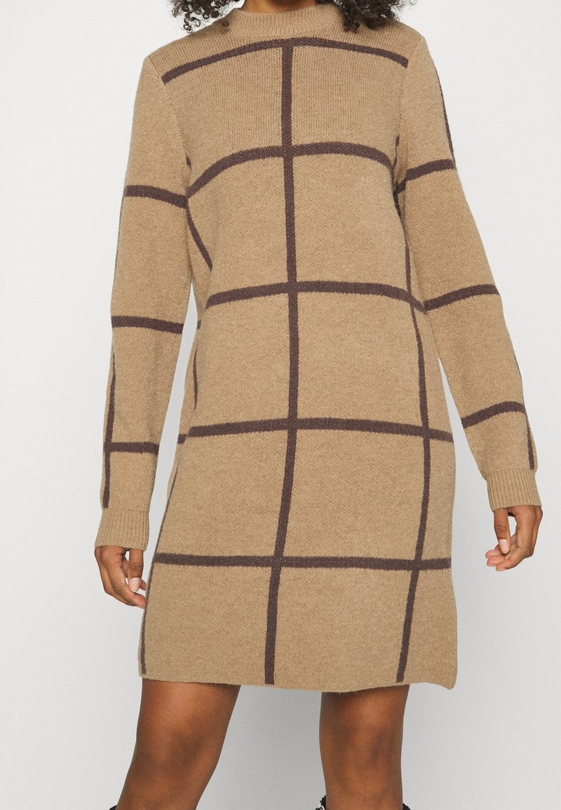 Robe en maille à manches longues à carreaux marron portée par une personne à la peau de ton moyen, visible du cou jusqu'au milieu des cuisses, sur un fond blanc.
