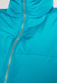 Veste matelassée bleu vif avec une fermeture éclair centrale argentée et un col montant, vue de face en gros plan.