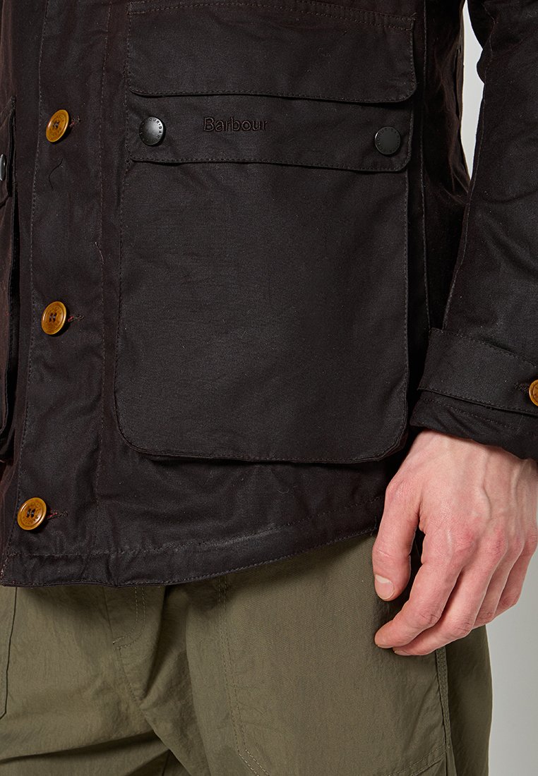 Close-up of dark brown Barbour jacket with large buttoned pocket, wooden buttons, and a person's hand resting beside olive green pants.