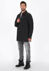 Mand iført sort frakke med brun krave, grå jeans og sorte støvler, stående mod hvid baggrund, holder frakken nær kraven.