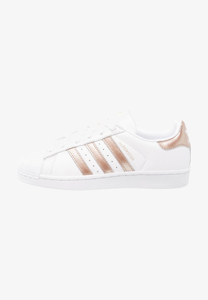 Zapatilla de cuero blanca con acentos en rosa dorado, diseño de tres franjas, puntera redondeada y suela de goma texturizada. Marca clásica en el lateral.