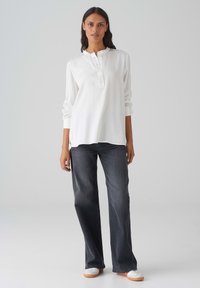 Camicia bianca a maniche lunghe con colletto henley e bottoni, abbinata a jeans svasati grigio scuro e sneakers bianche con suole arancioni.