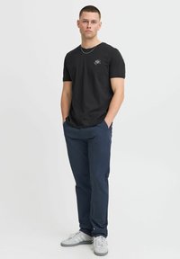 Camiseta de algodón negra con un pequeño diseño bordado en blanco, combinada con pantalones azul marino oscuros y zapatillas gris claro. Diseño simple y ajustado.