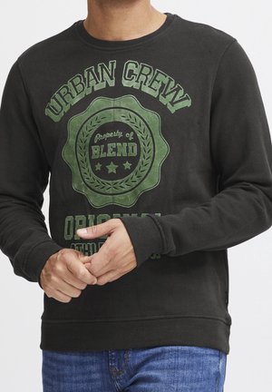 Mężczyzna w czarnej bluzie z kapturem z zielonym emblematem "Urban Crew Property of Blend" i niebieskich dżinsach, poprawiający rękaw obiema rękami.