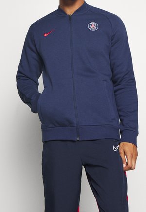 Mand iført marineblå jakke med fuld lynlås med Paris Saint-Germain-logo og rød Nike-swoosh, marineblå bukser med hvidt Nike-logo.