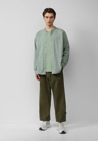 Lichtgroene button-down overhemd over een bijpassend groen T-shirt, gecombineerd met losse olijfgroene broeken en witte sneakers met grijze accenten.